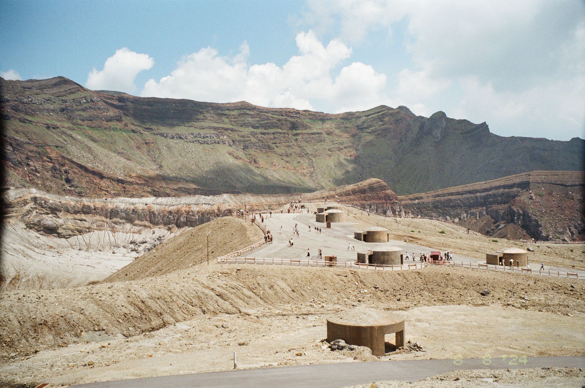 Tourists walking around the scenic crater area of Mount Aso, Japan, under a bright blue sky.