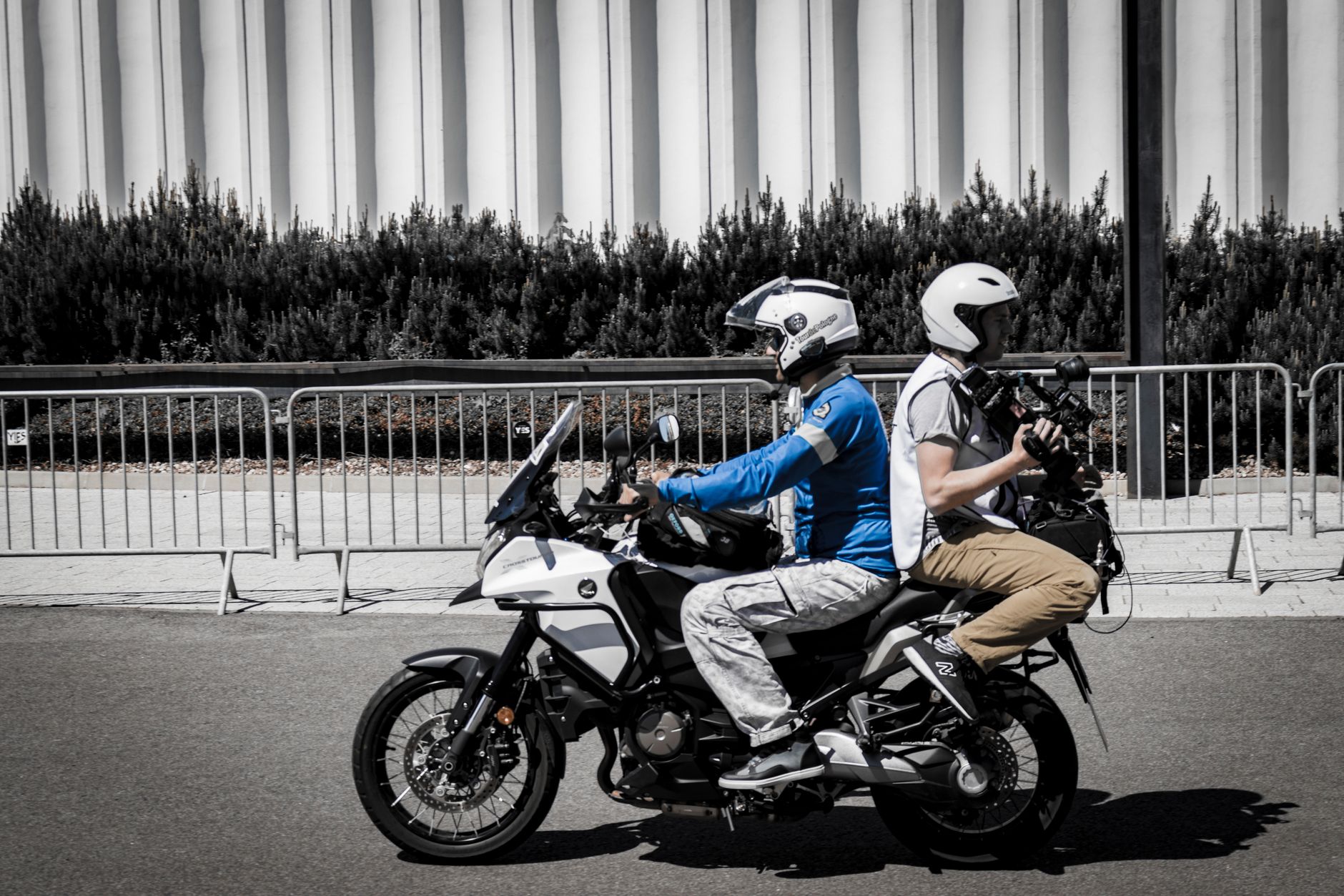 Two motorcyclists capturing video on a street with modern architecture.