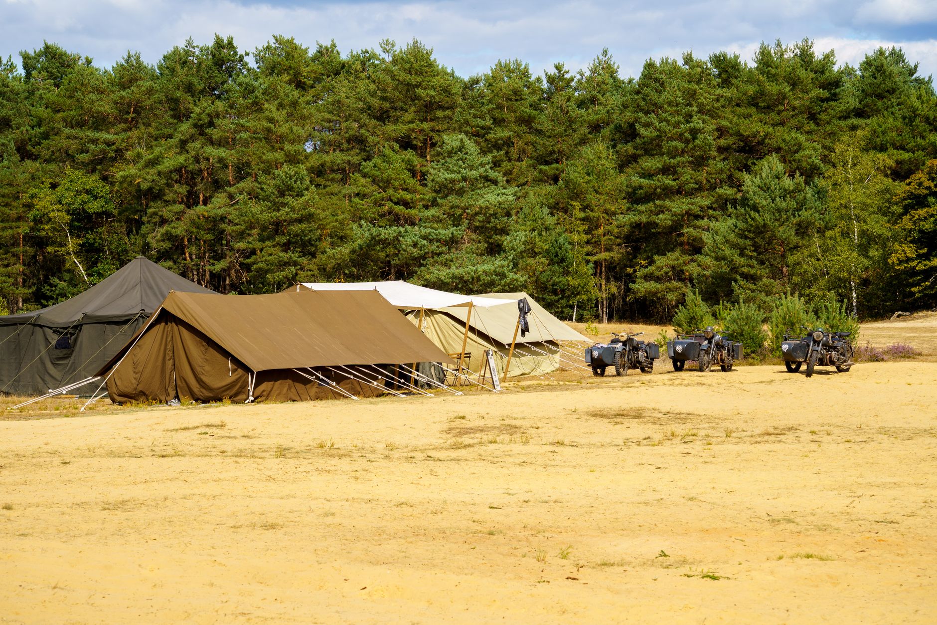 Outdoor military camp in a sandy area with tents and vintage motorcycles against a forest background.