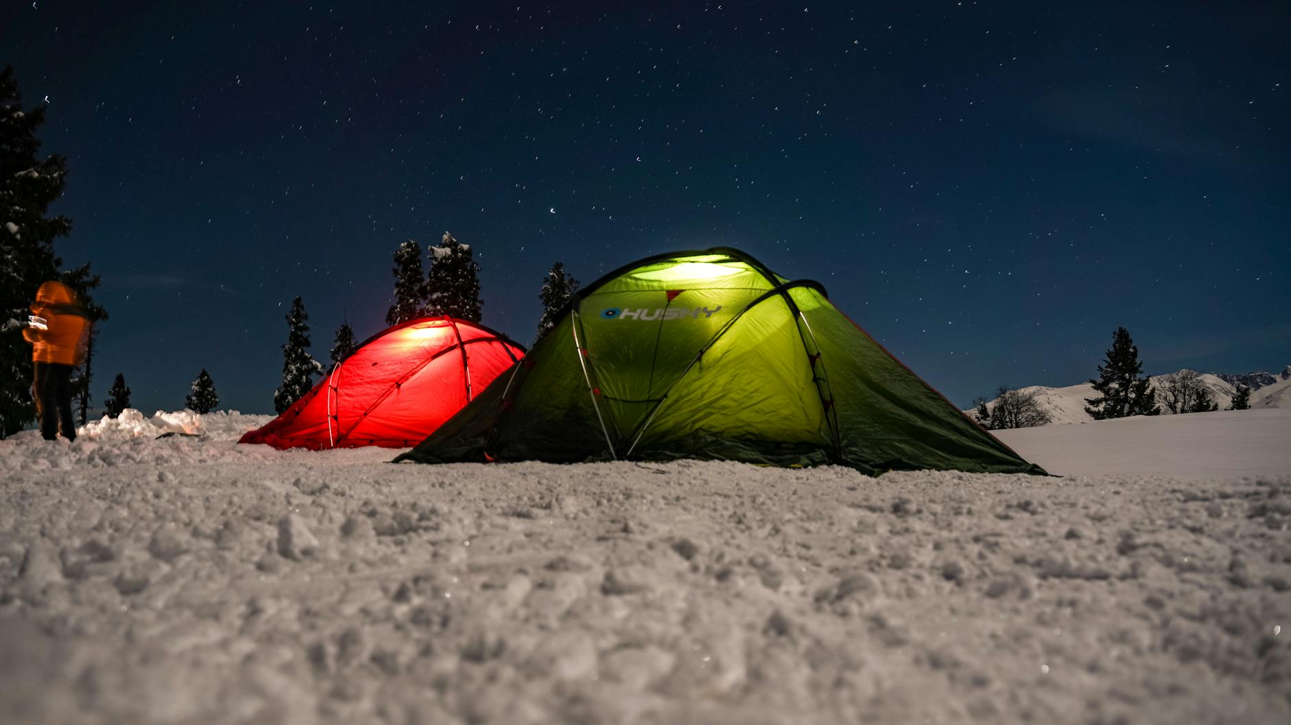 Two tents illuminated at night, set on a snowy landscape beneath a clear starry sky.