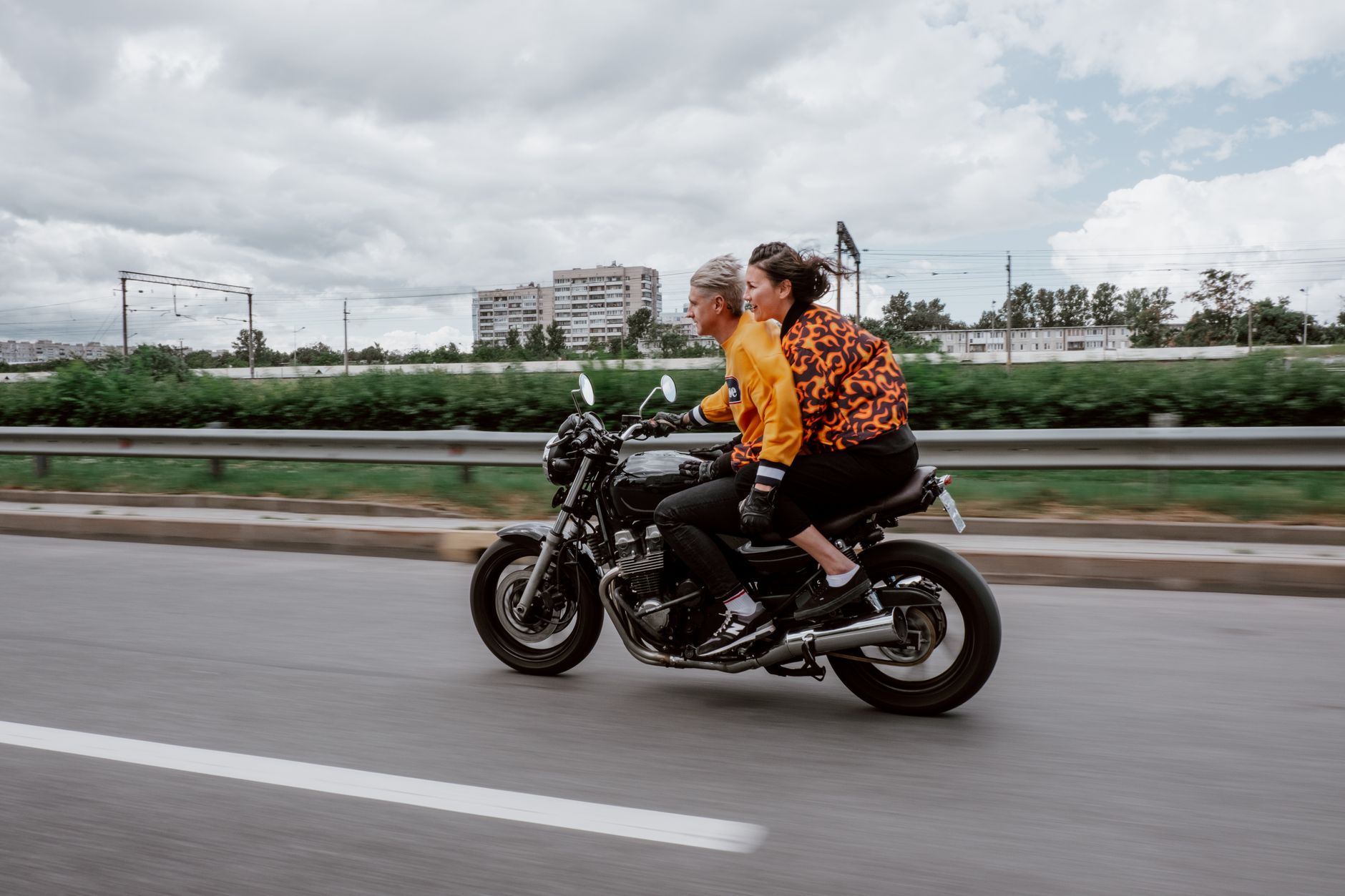 A couple enjoys a thrilling ride on a motorcycle along a highway lined with greenery and buildings.
