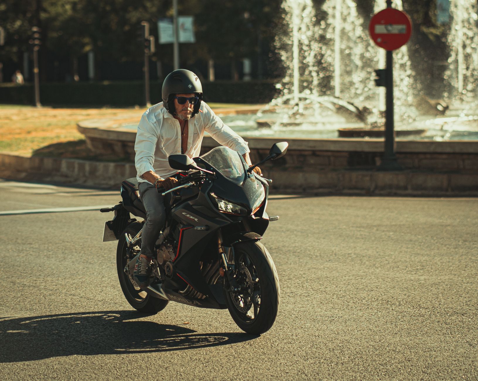 A man rides a motorcycle wearing a helmet near a scenic fountain in a city street.