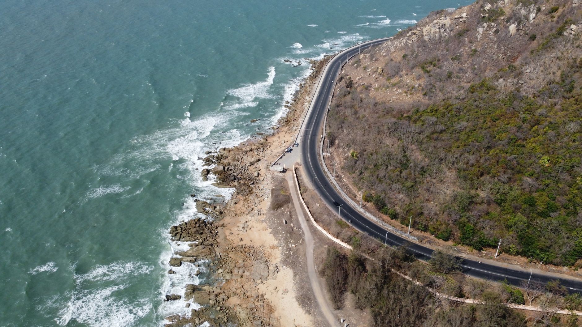 A breathtaking view of a winding coastal road along a rocky shoreline with waves crashing against the rocks.