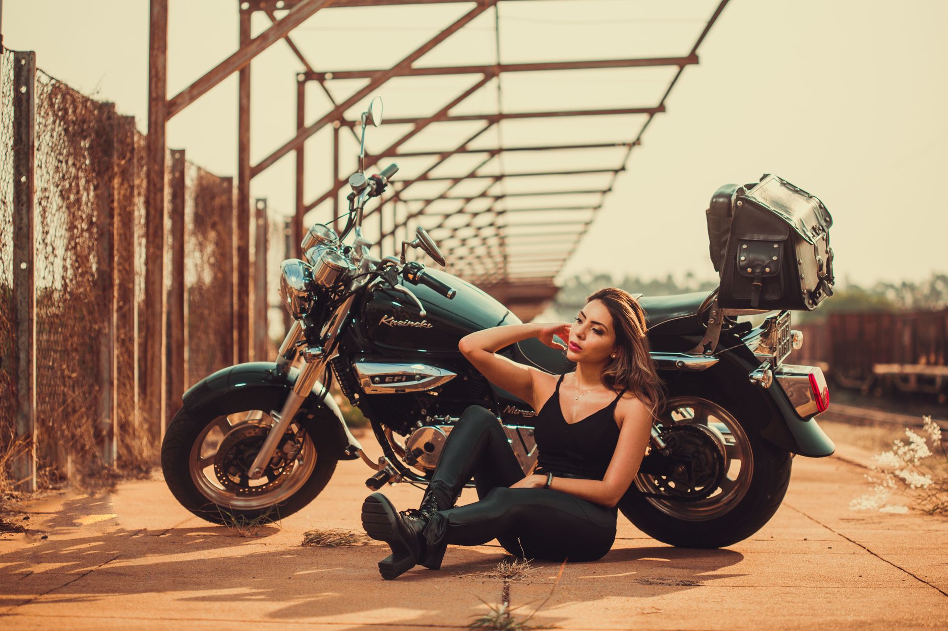 A fashionable woman in black outfit sitting by a parked motorcycle on a sunny day.
