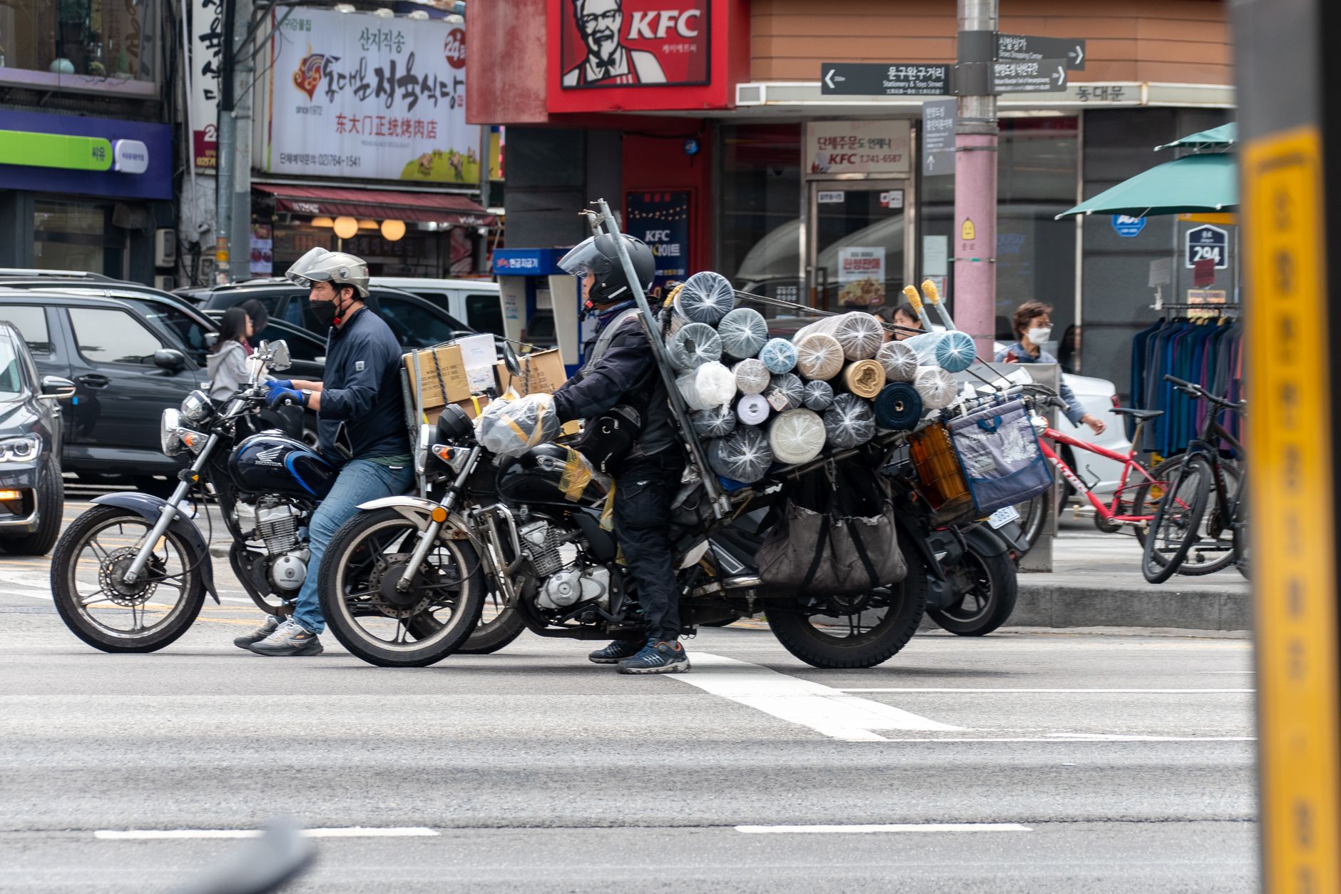 Motorcyclists carrying large cargo loads wait at a city intersection near recognizable fast-food outlets.