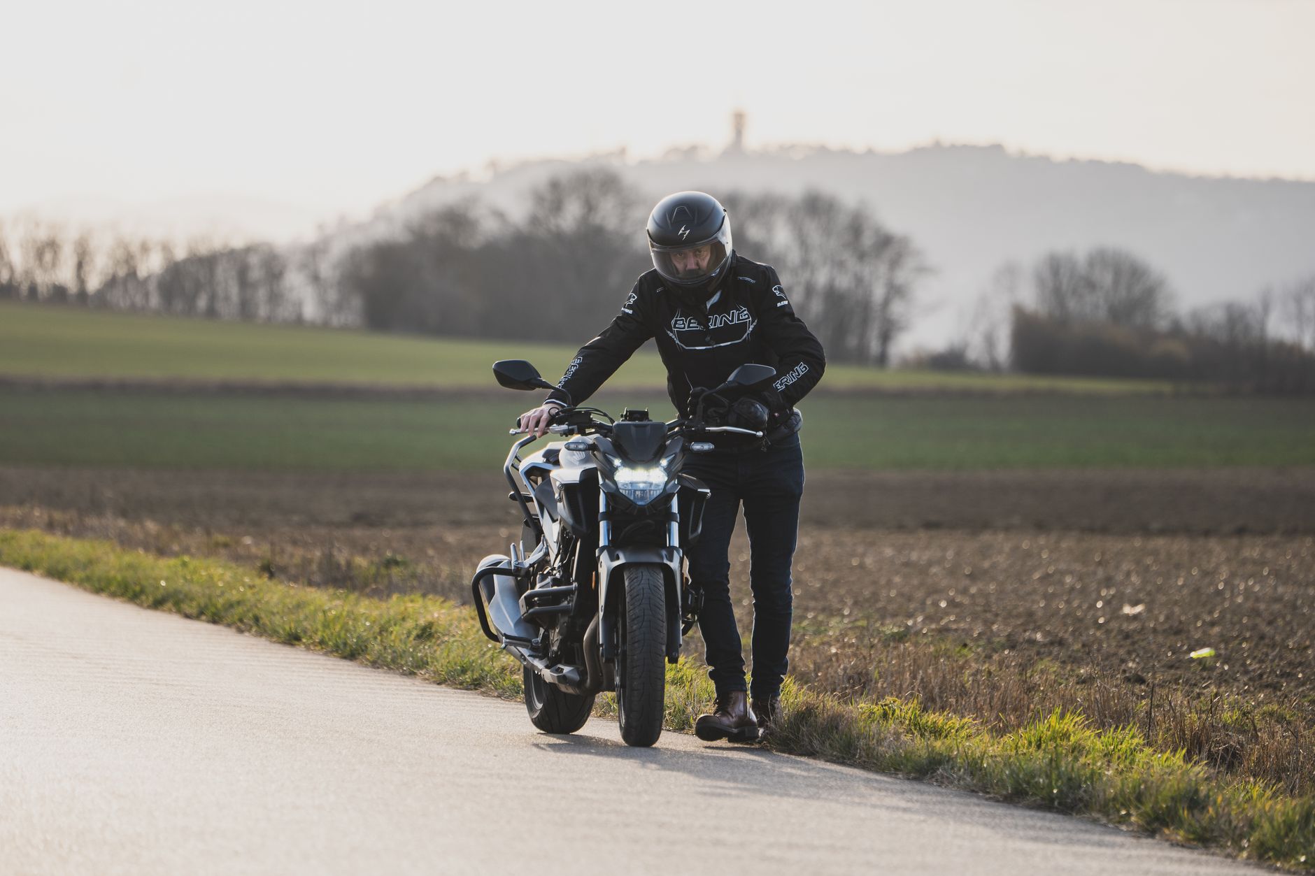 A biker stands next to a motorcycle on a rural roadside, helmet on.