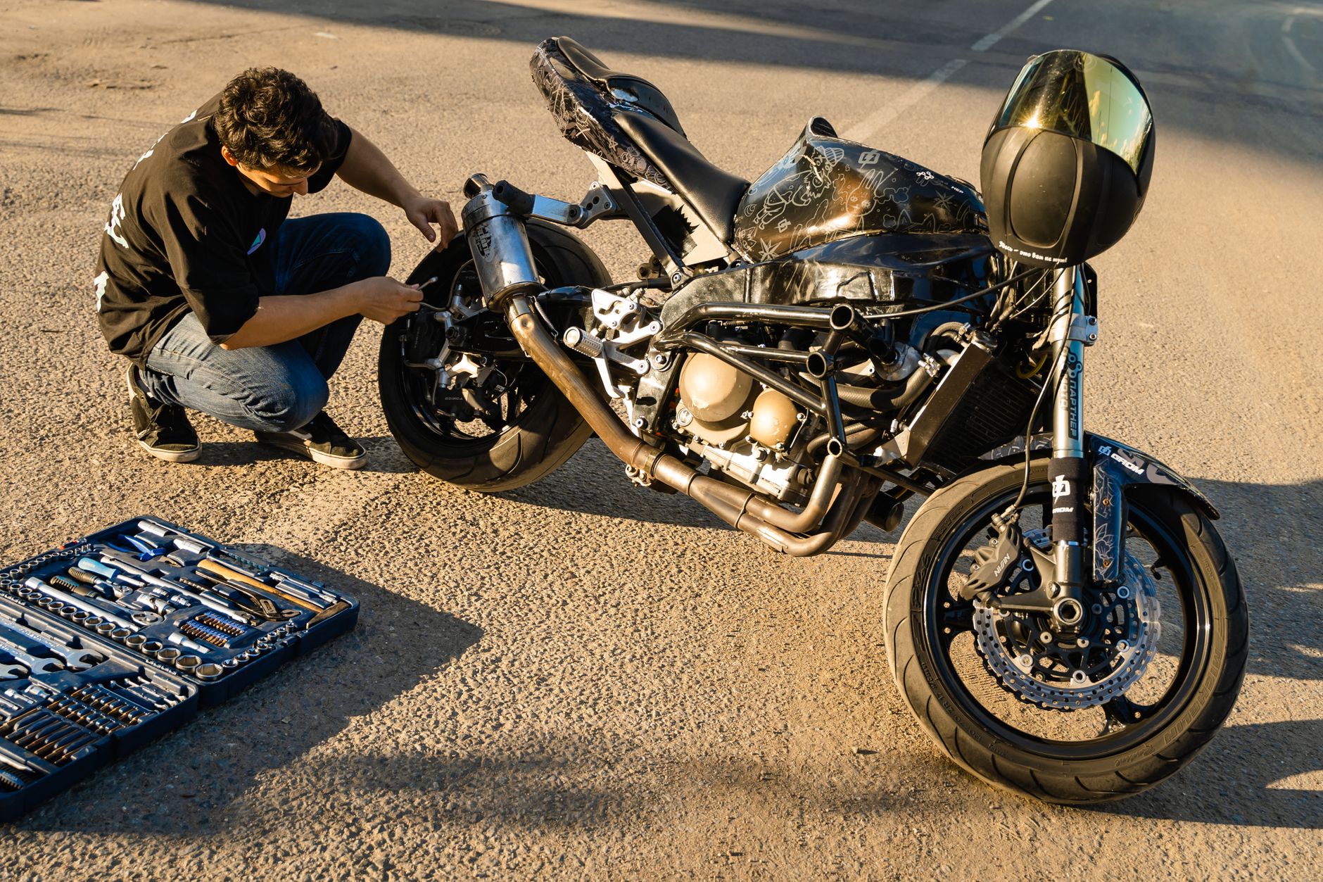 A man in a black shirt adjusts a motorcycle on a sunlit concrete surface with a toolbox nearby.