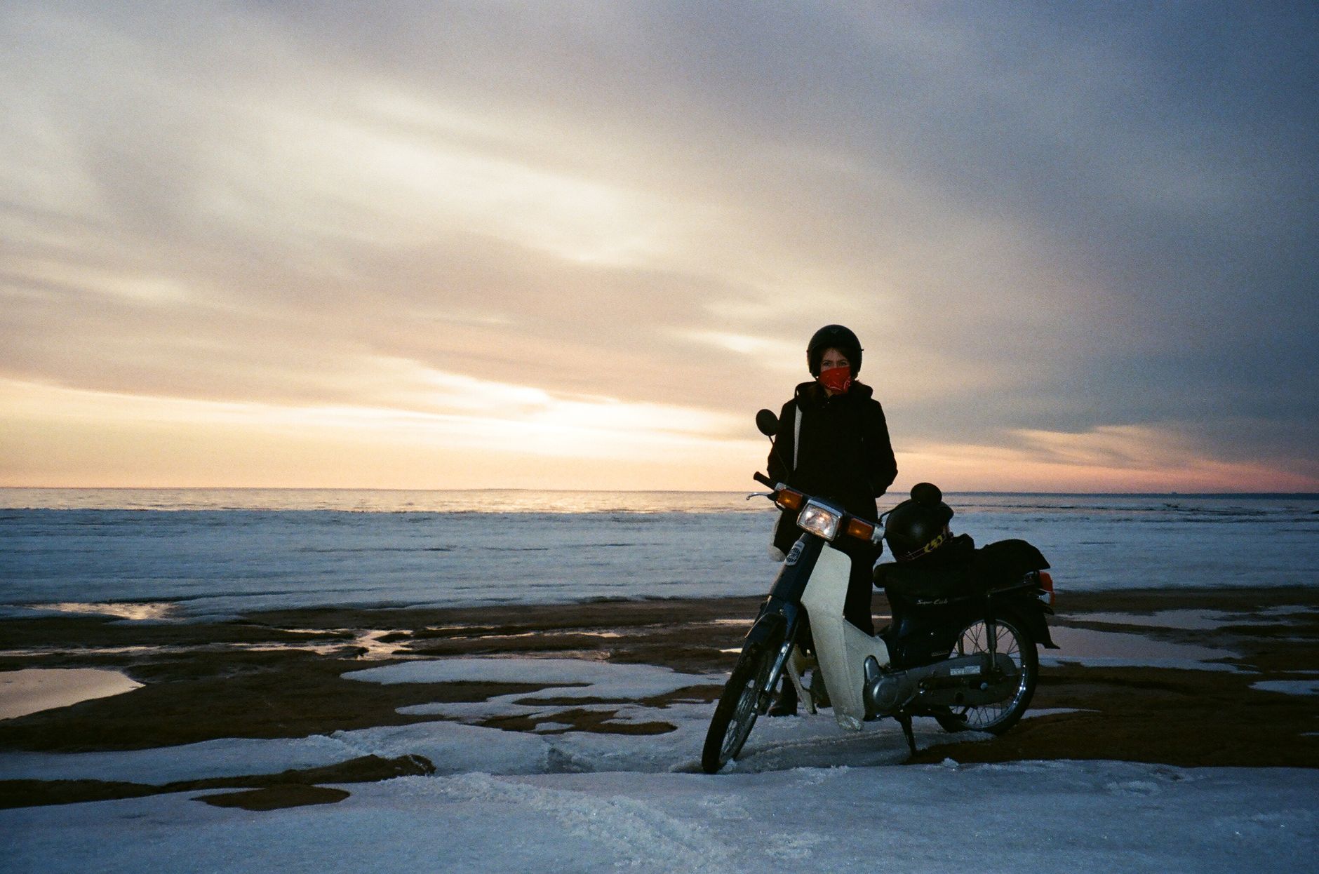 A woman on a motorcycle by a snowy seashore during twilight, offering a serene winter scene.