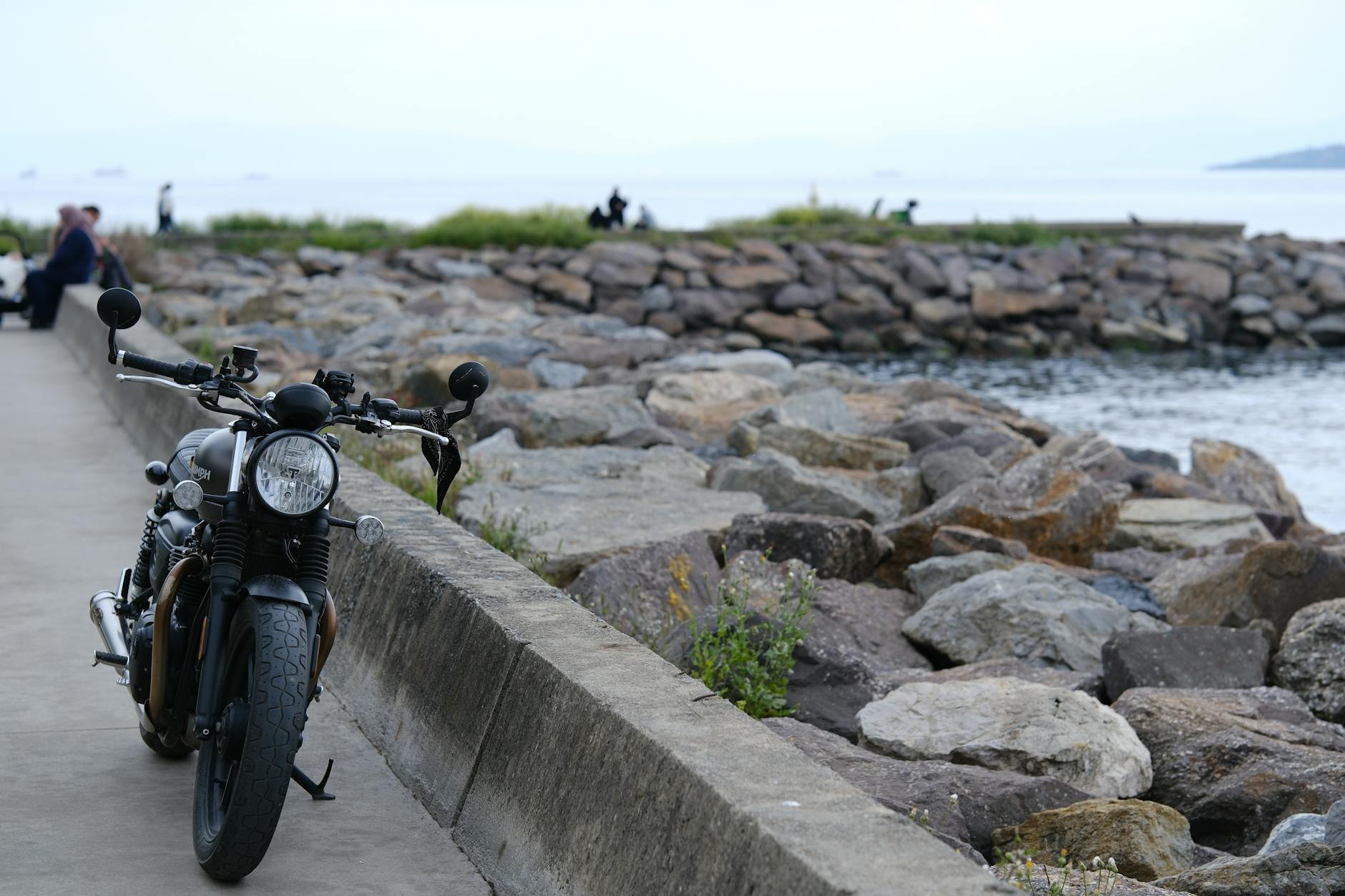 A classic motorcycle parked on a coastal path with rocks and the sea visible in the background.