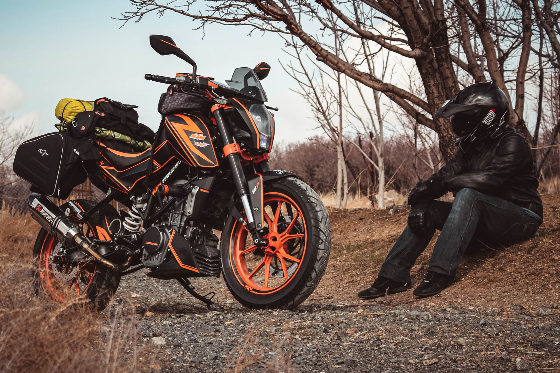A motorcyclist in gear rests beside a packed KTM bike on a rural path.