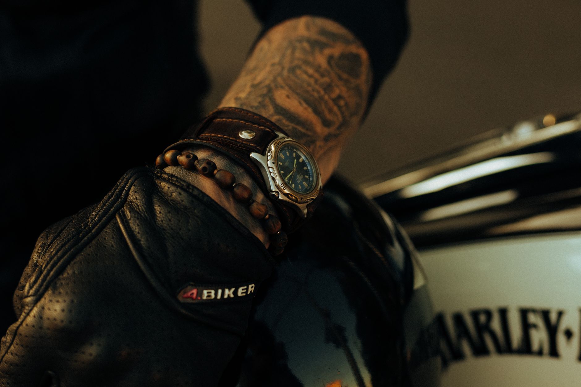 A masculine arm with tattoos wearing a watch and gloves, leaning on a motorcycle.