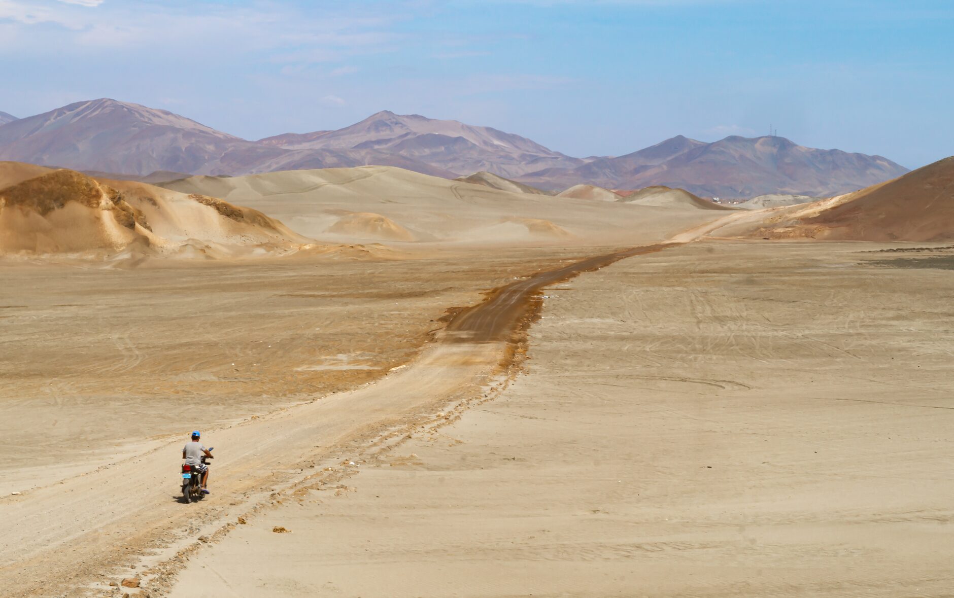 A lone motorcyclist traverses the sandy dunes of Huarmey, Peru, under a bright summer sky.