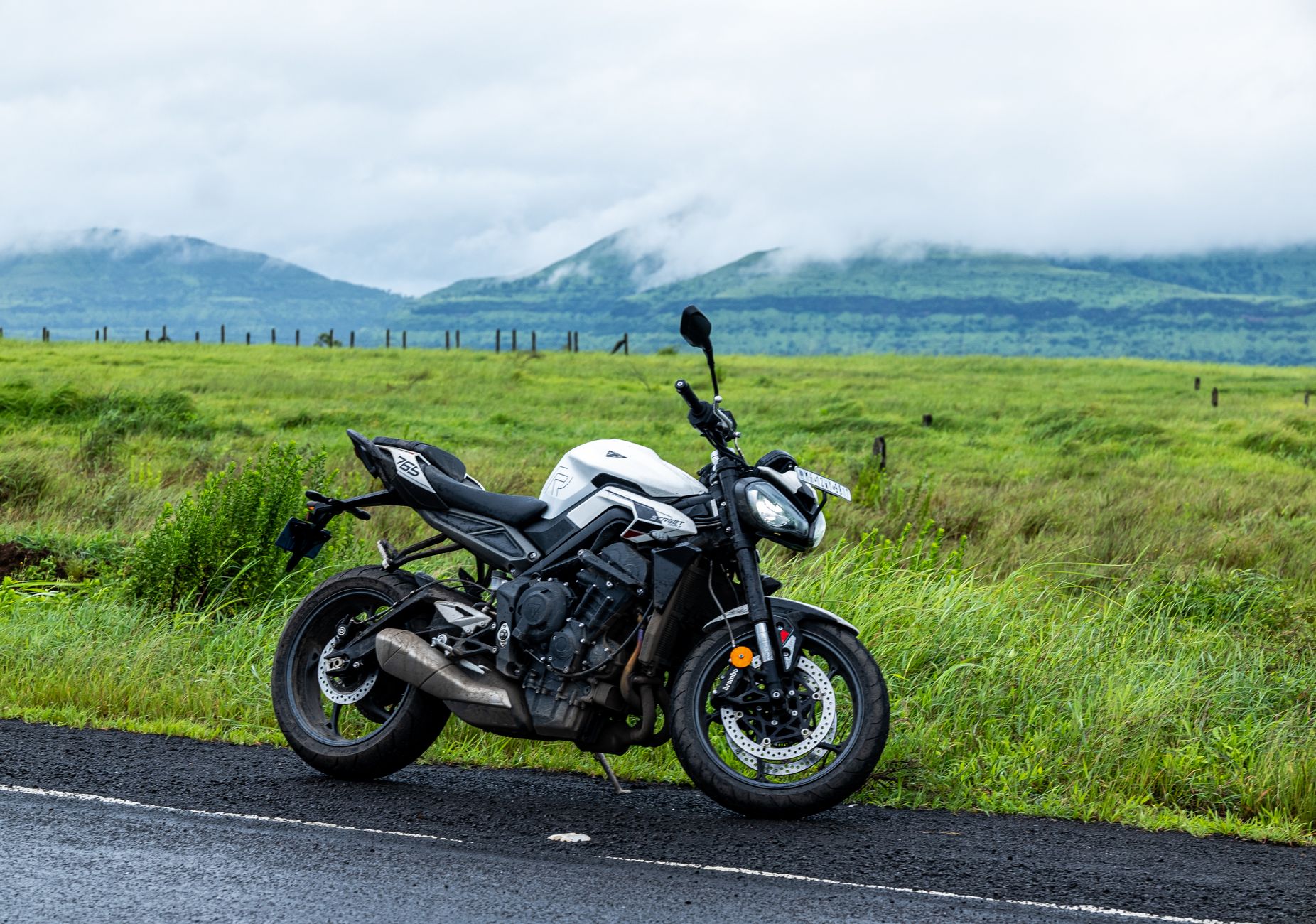A motorcycle parked on a road with lush green fields and misty mountains in Kas, MH, India.