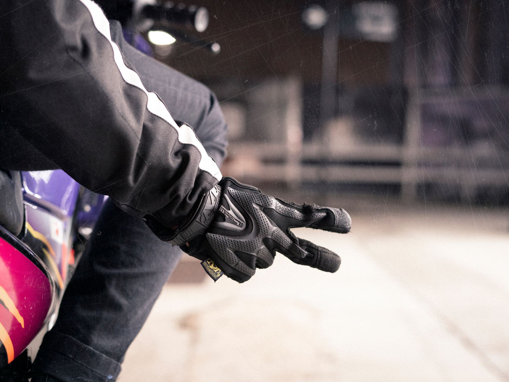 Close-up of a motorcyclist's gloved hand showing peace sign, with blurred urban background.