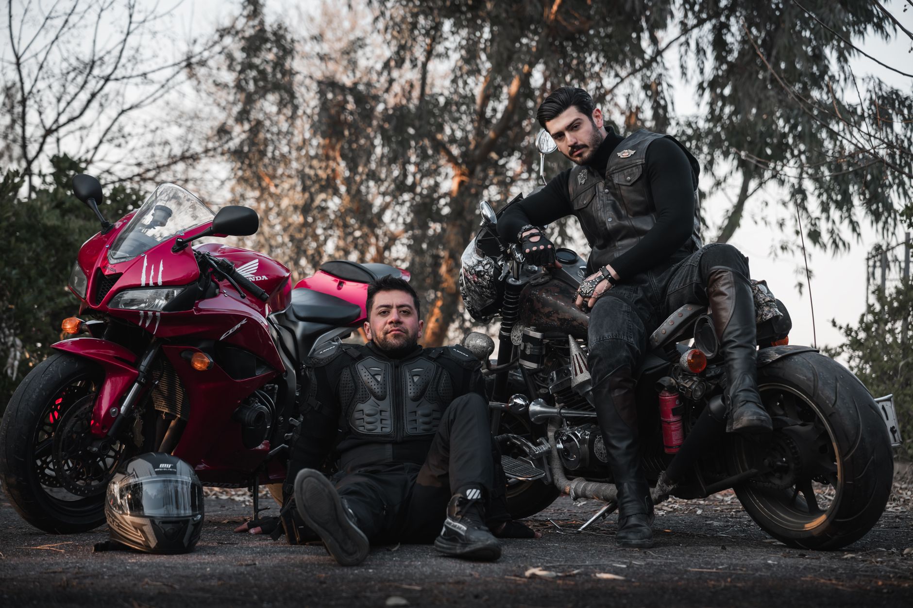 Two bikers in gear pose confidently with motorcycles on an outdoor setting.
