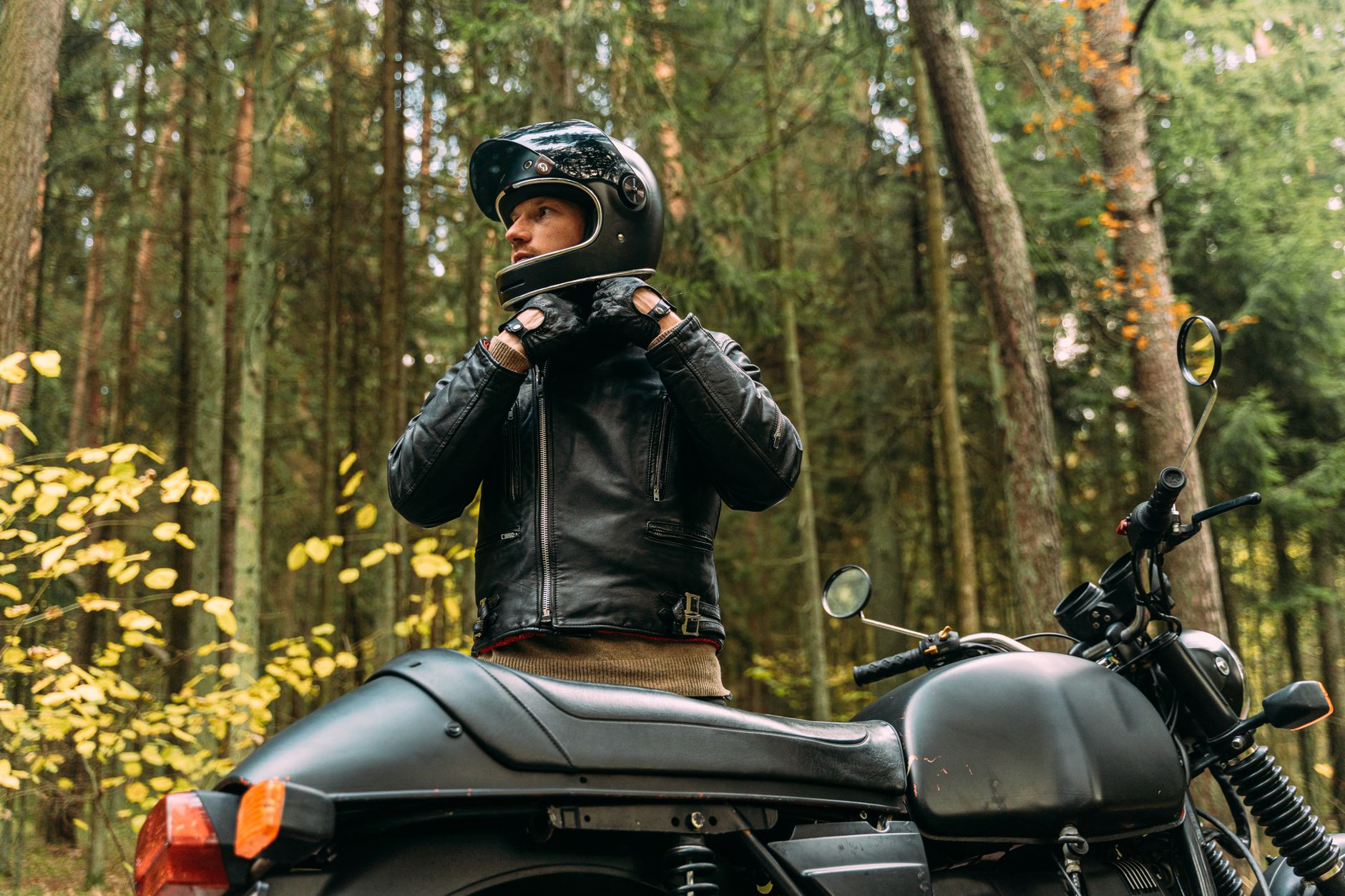 A man in black leather gear prepares for a motorcycle journey in a forest setting, embodying adventure and freedom.