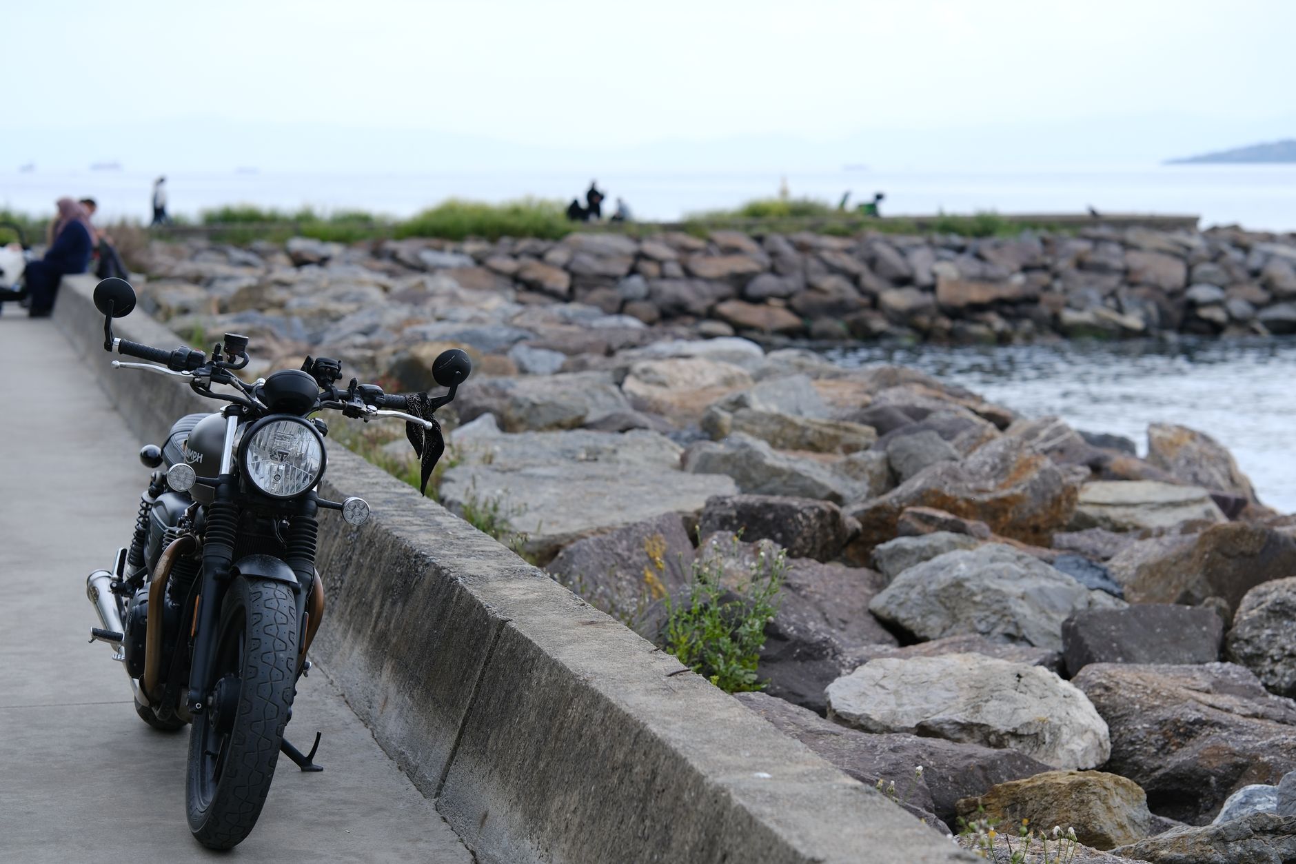 A classic motorcycle parked on a coastal path with rocks and the sea visible in the background.