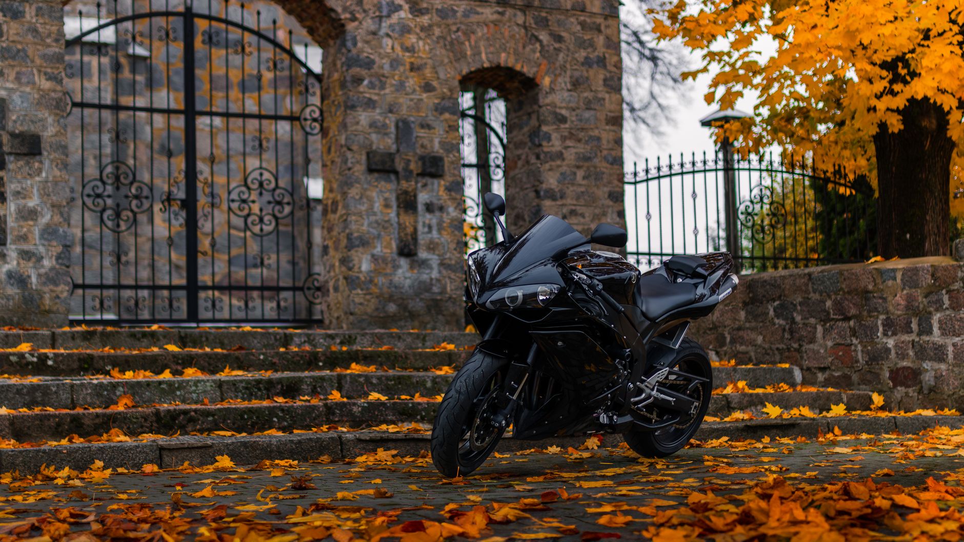 Black motorcycle parked on stone steps surrounded by autumn leaves and a historic gate.