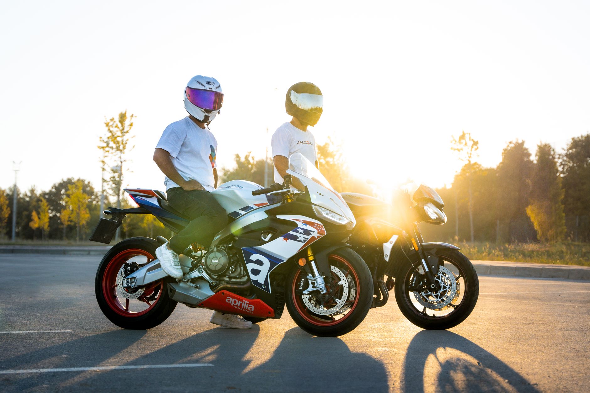 Two bikers with motorcycles standing in a sunny parking lot, helmets on, during sunset.